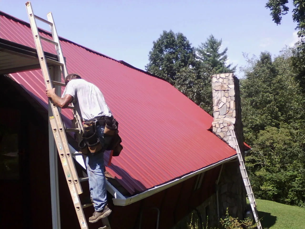 Roofer on a ladder beside a red metal roof and stone chimney — Soffit & Fascia Repair by Roofing Brothers in Sylvania